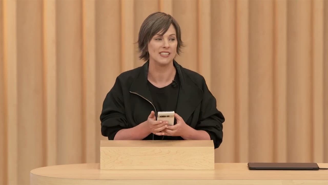 Cathy Edwards em palestra durante evento do Google. Ela é uma mulher de cabelos curtos e morenos, penteados para o lado, e está usando camisa e jaqueta preta. Ela segura um celular branco nas mãos e fala em um púlpito em um palco. 