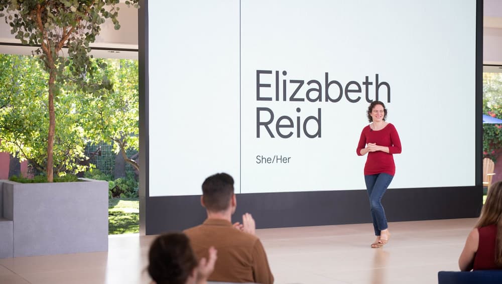 uma mulher de calça jeans e blusa vermelha caminha em frente da um telão branco que ocupa quase toda a parede, onde se lê "Elizabeth Reid, she/her"